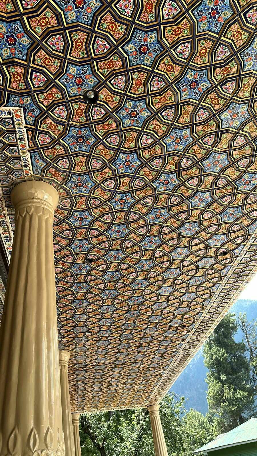A hand-painted khatamband ceiling seen from below in a Kashmiri pavilion, gold and lapis star panels running away over turned columns.