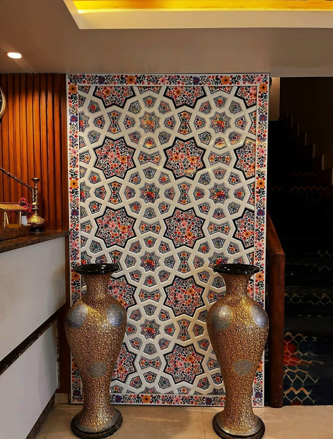 Khatamband wall panel of ivory star interlace with painted flowers behind a hotel reception desk, flanked by large gold paper machie vases.